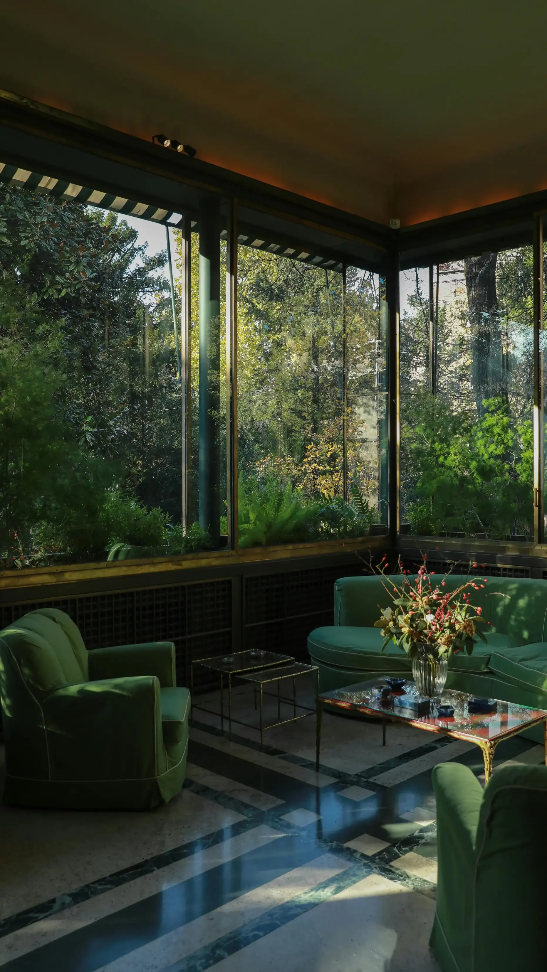 Glass-enclosed sunroom with green velvet seating and lush garden views