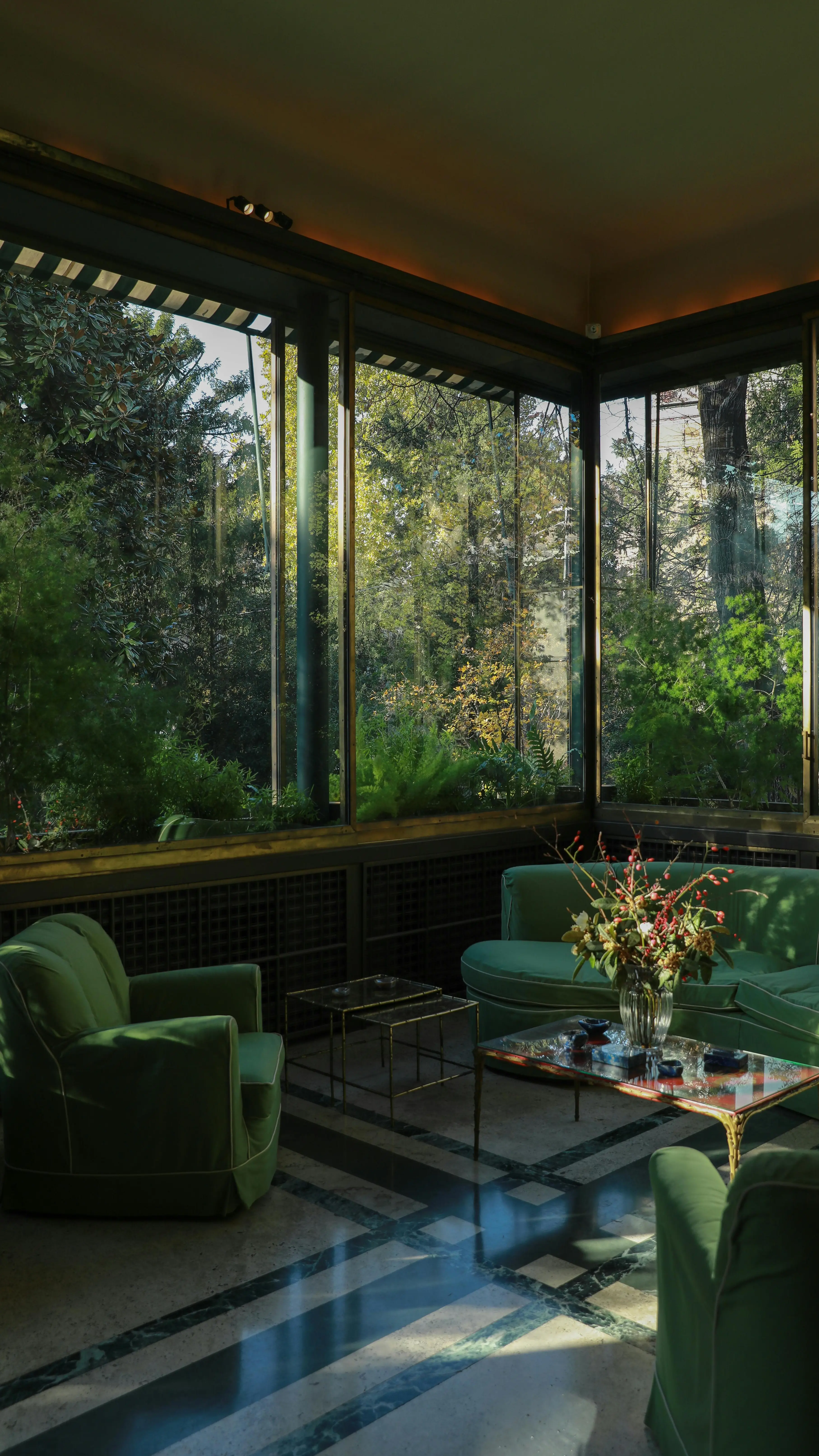Elegant glass-enclosed sunroom with emerald green velvet seating, gold coffee table and lush garden views